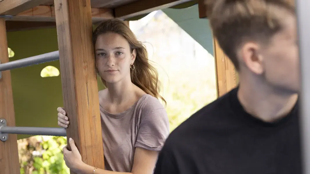 Unscharf im Vordergrund: Abgewandter Junge, nach links blickend. Versetzt dahinter: zugewandtes Mädchen, Blick in die Kamera gerichtet