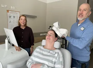 Oberarzt Dr. Lars Bretz (rechts) demonstriert an der Fachangestellten Rebekka Herrmann (Mitte), wo man das Gerät am Kopf positioniert, damit es optimal arbeitet. Klinikdirektorin Dr. Stefanie von Rosen (links) freut sich, ihren Patientinnen und Patienten diese weitere Therapiemöglichkeit anbieten zu können.