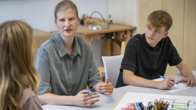 Selbsthilfe will gelernt sein Junge Frau sitzt zwischen zwei jugendlichen Kindern am Tisch. Ein Kind blickt aufmerksam zur Frau, beide halten eine Stift in der Hand.