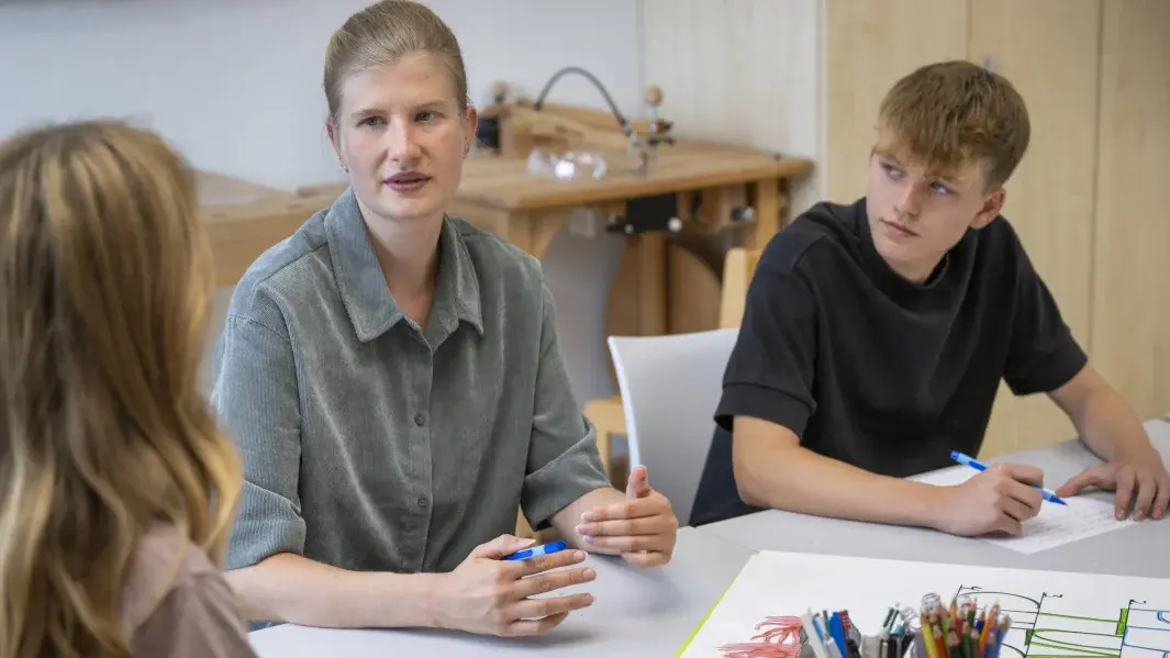 Junge Frau sitzt zwischen zwei jugendlichen Kindern am Tisch. Ein Kind blickt aufmerksam zur Frau, beide halten eine Stift in der Hand.