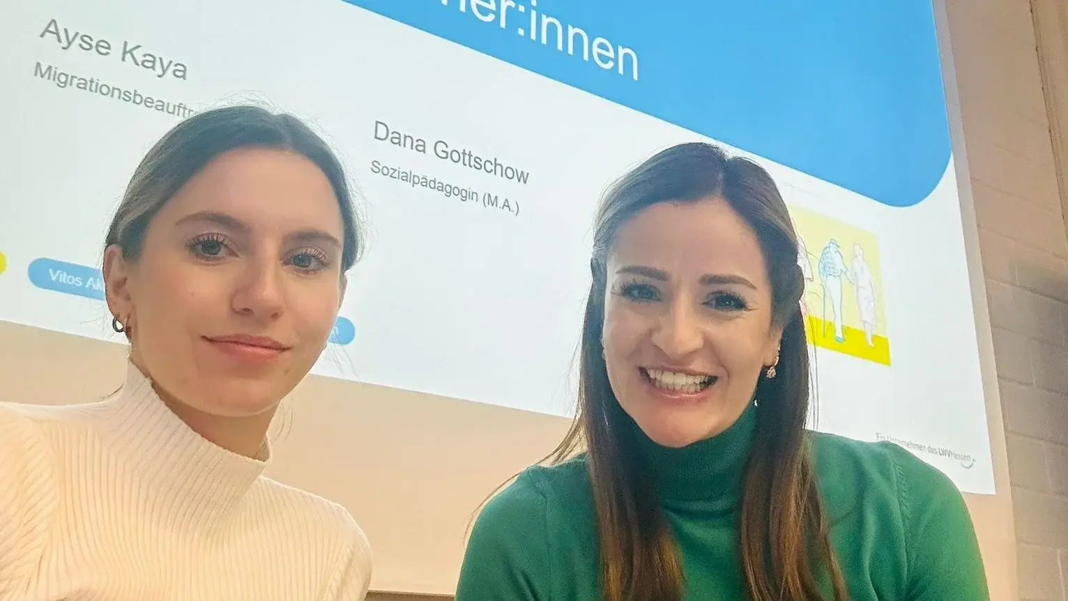 Zwei junge Frauen lächeln in die Kamera, im Hintergrund ist eine Präsentation zu sehen